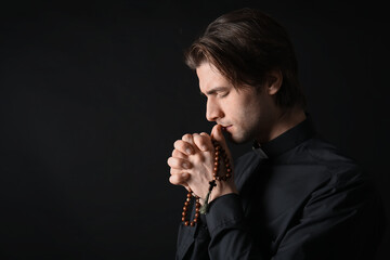 Young priest praying with beads on dark background, closeup