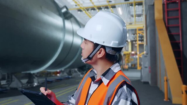 Close Up Side View Of Asian Male Engineer With Safety Helmet Standing In Pipe Manufacturing Factory. Taking Note On The Tablet And Looking Around