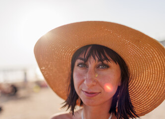 Close-up of the face of an attractive girl wearing a straw hat. Portrait of a happy young woman in a summer hat with a large brim on the beach. - 784927055
