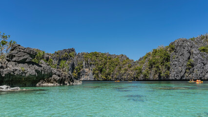 Obraz premium Tourist canoes sail through an amazing turquoise lagoon surrounded by picturesque karst rocks. Green vegetation on steep slopes. Clear blue sky. Philippines. Big Lagoon. Miniloc Island. Bacuit bay