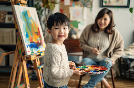 A Chinese child is drawing on an easel with his mother next to him, holding colorful paint palette and brush in hand