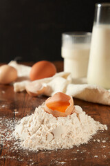 A cracked egg with shells in a pile of flour on wooden kitchen table with some baking ingredients blurred behinds. Cooking concept for product make from eggs or pastry, front view photo