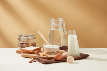 Front view of a group of baking ingredient with water jug, a milk bottle, a jar of nut, sandwiches, eggs and flour, decorated by brown towel and chopping board. 