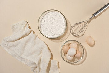 An array of baking ingredients and utensils neatly arranged on pink counter top, including flour, eggs, and whisk, preparation for cooking. Top view photo, space for adding design elements