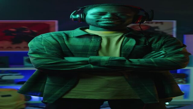 Vertical zoom-in medium portrait of African American gen z guy wearing checkered shirt and gaming headphones posing for camera with hands folded standing in dark room with neon light