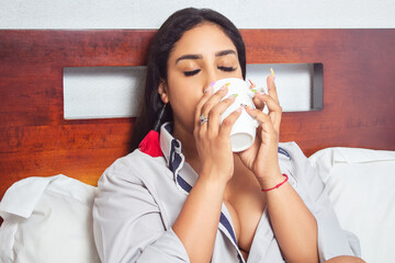 portrait of beautiful and sensual mexican latin woman drinking cup of coffee, wearing shirt sitting on bed