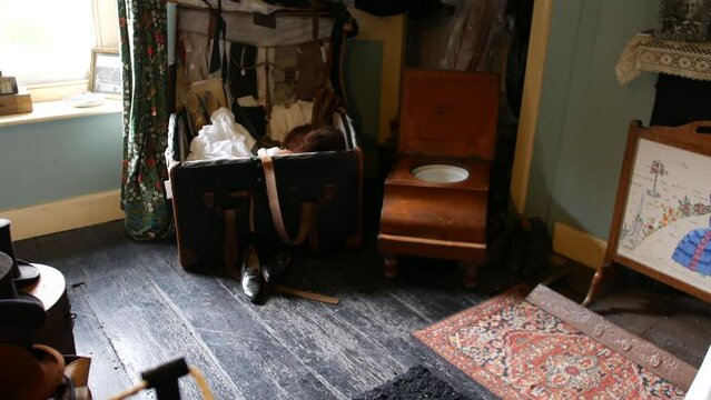 Vintage room with old-fashioned luggage, wooden commode chair, and patterned textiles, evoking historical home life. Antique ambiance with worn floor and classic decor for period films