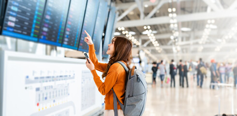 Young asian woman in international airport, using mobile smartphone and checking flight at the flight information board