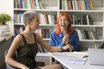 Two girls students sit at desk in high-school library, talking, laughing, enjoy pleasant...