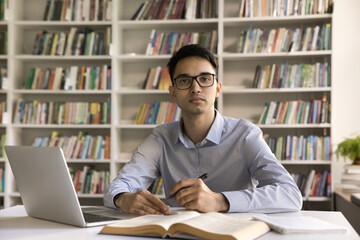 Portrait of serious diligent Indian student guy in glasses make exercise, writes summary studying using laptop seated at desk with textbooks and copybook, look at camera. Higher education, development