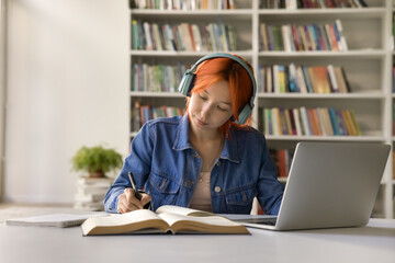 Focused red haired 17s student girl doing schoolwork, listen audio course improving foreign...