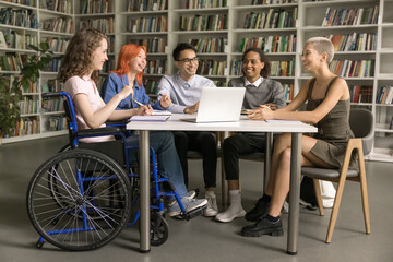 Pretty student girl with disability sits in wheelchair talking to interracial schoolmates, studying...
