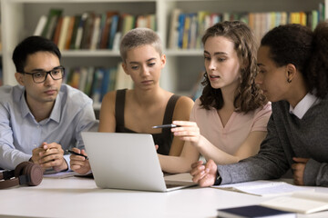 Four focused multiethnic students do homework together sit in library, discuss online task use laptop, prepare for exams, search information on internet, share ideas, work on group project. Education