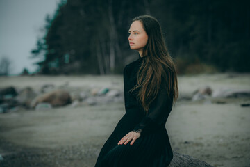 A woman is sitting on a rocky shore with waves crashing behind her. Black dress, checkered coat....
