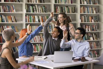 Happy interracial students giving a high five gesture greeting or encouraging each before exams seated at desk in classroom on bookshelves background. Unity, support, companionship, educational event - obrazy, fototapety, plakaty