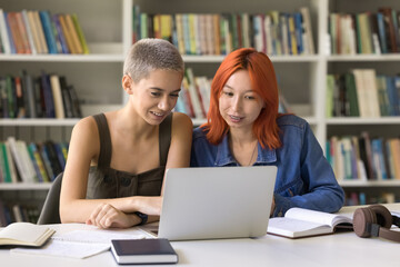 Two attractive girls student watching educational video on laptop, doing homework together, communicate, gain new knowledge, prepare for exams, read lecture on internet, studying in university library
