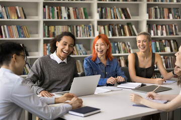 Interracial group of young students met in library sit at table laughing on jokes, distracted from...