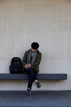 Young Chinese student sitting on bench at university campus with mobile phone