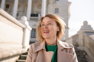 A woman with long hair is standing on a set of stairs. She is wearing a green shirt and a tan coat.
