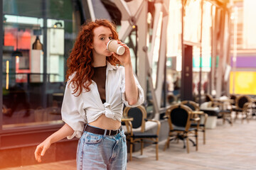 happy young redhead woman in jeans and white shirt drinking take away coffee on street and having...