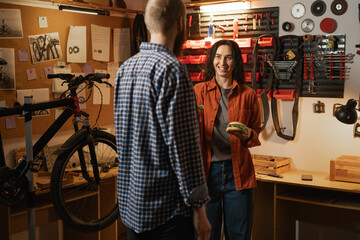 Couple bicycle mechanic in repair workshop. In cycle garage man and woman eating burgers on lunch....