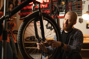 Young beard bicycle mechanic repairing bicycles in a workshop or garage, fixing bicycle wheel.