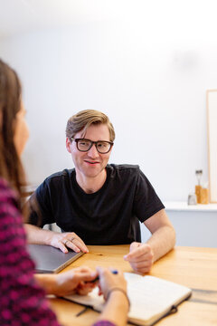 Caucasian man wearing thick rimmed glasses sitting across from a woman at a table in an office