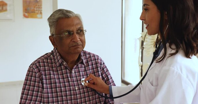 Happy young Latin doctor woman examining older patient, listening to heartbeat with stethoscope, checking pulse rate, heart work, breathing, smiling, talking to senior Indian patient in exam room