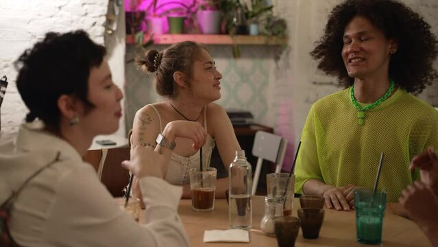 Homosexual friends have fun at table with alcohol in bar. Young lesbian women, gay man talking with smiles and laughing, drinking cocktails and sitting in room. Concept: non-binary multiethnic people