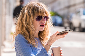 middle aged woman sending a voice or audio message with the smartphone and drinking a takeaway on...