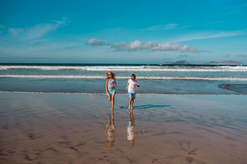Siblings playing on beach, running, skipping in water. Smilling girl and boy on sandy beach of Canary islands. Concept of family beach summer vacation with kids.
