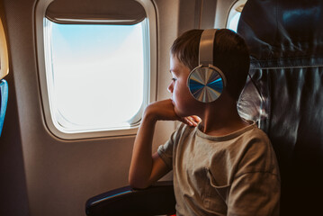 Boy with headphones sitting airplane, looking out of window. Concept of family beach summer vacation with kids.