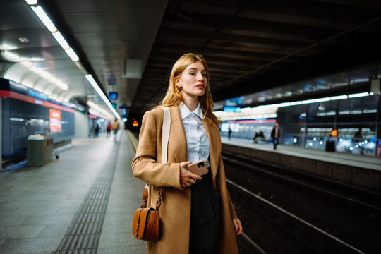 Female standing at subway platform with mobile phone, using social media and waiting train