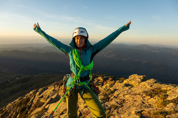 A woman is standing on a mountain top, wearing a green harness and a helmet. She is smiling and she...