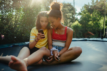 Teenager girls friends spending time outdoors in garden, laughing. Sitting on trampoline, scrolling on smartphone, social media.