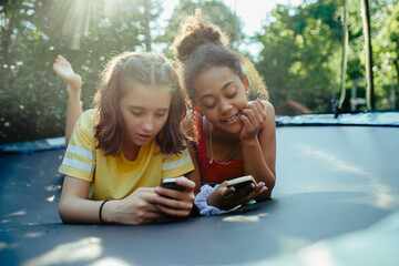 Teenager girls friends spending time outdoors in garden, laughing. Lying on trampoline, scrolling on smartphone, social media.