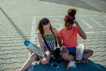 Teenager girl best friends with skateboards spending time outdoors in city during warm summer holiday day. Sitting on parking lot, talking.