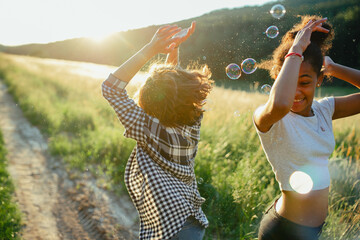 Young teenager girl best friends spending time in nature, during sunset. Girls blowing bubbles and popping them.