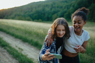 Young teenager girl best friends spending time in nature, during sunset. Girls on walk, taking selfie with smartphone. - obrazy, fototapety, plakaty