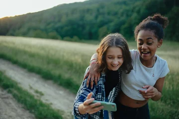Schilderijen op glas Young teenager girl best friends spending time in nature, during sunset. Girls on walk, taking selfie with smartphone. © Halfpoint