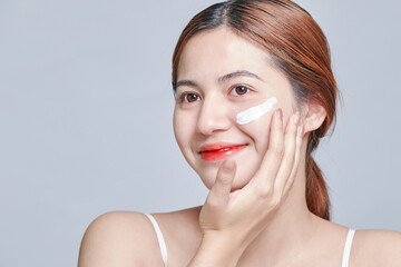 Positive young female in white top applying moisturizing cream on face. Close up beauty portrait of...