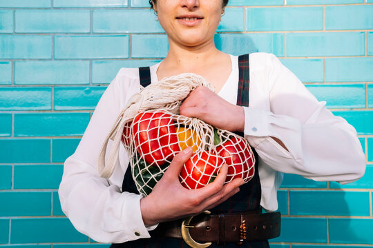 Woman holding bell peppers in mesh bag near tiled wall