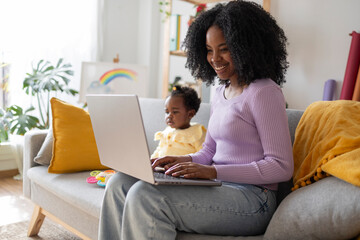 Smiling young mother using laptop working at home sitting by daughter on sofa in living room