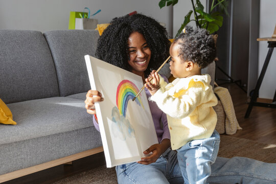Happy mother holding canvas for baby girl painting rainbow with paintbrush at home