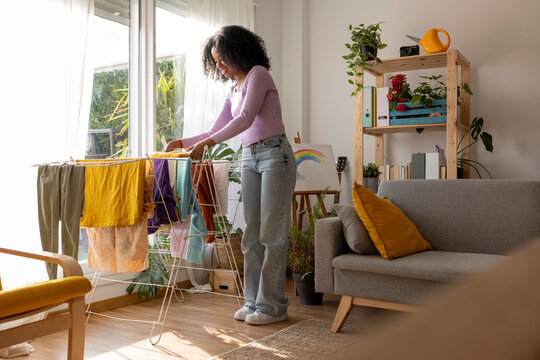 Smiling young woman drying clothes on rack by sofa in living room