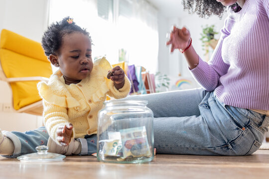 Young mother sitting near daughter playing with coins by jar of currency at home