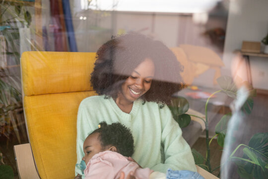 Smiling mother sitting on chair with baby girl seen through window glass