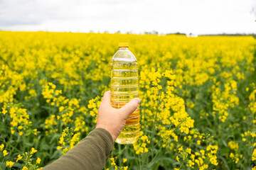 A bottle of rapeseed oil in a hand against the background of a yellow blooming rapeseed field. A bottle of rapeseed oil and blooming rapeseed fields. - obrazy, fototapety, plakaty