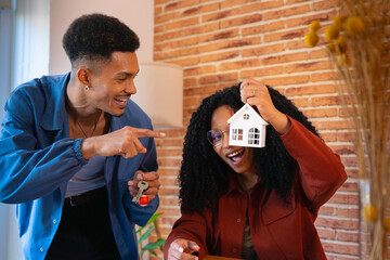Happy multiracial couple showing a mini wooden house and a key with a key ring. 
