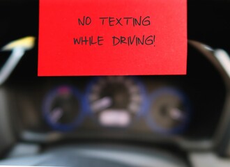 Red sticky note with text written  NO TEXTING WHILE DRIVING  on the car steering wheel , to remind...